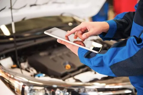 A mechanic in overalls using a tablet while inspecting a vehicle's engine bay