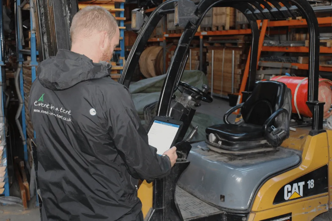 A warehouse worker completing a digital checklist on a tablet beside a forklift